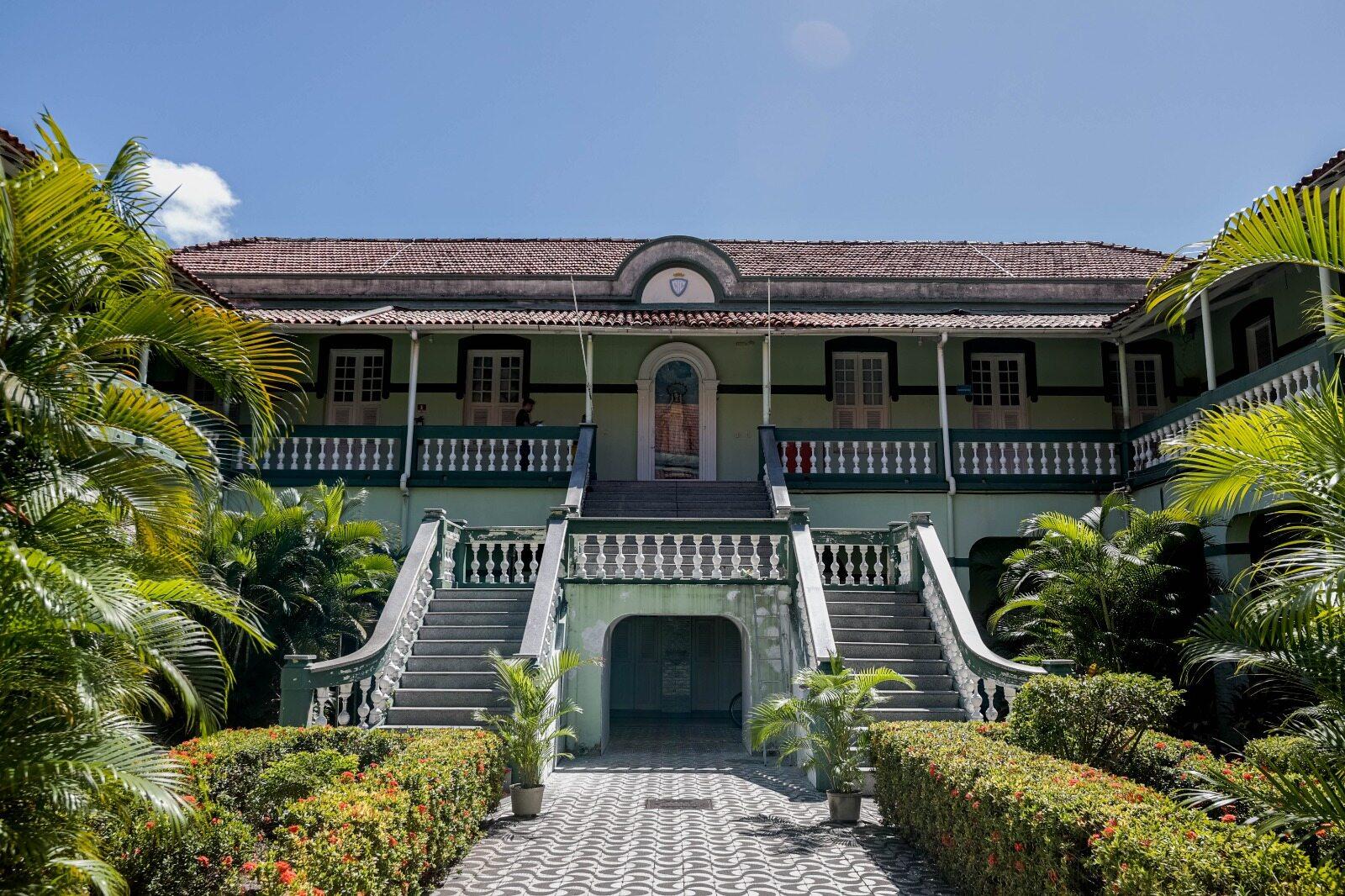A imagem mostra um casarão de dois andares com uma fachada em tom verde-claro e telhas de cerâmica. Duas escadas curvas levam ao andar superior. Há balaustradas brancas nos dois andares. A entrada principal tem um arco e porta de madeira. A frente da construção possui um pátio com caminhos de pedra, canteiros de flores, arbustos aparados e palmeiras. O céu está azul e com poucas nuvens.