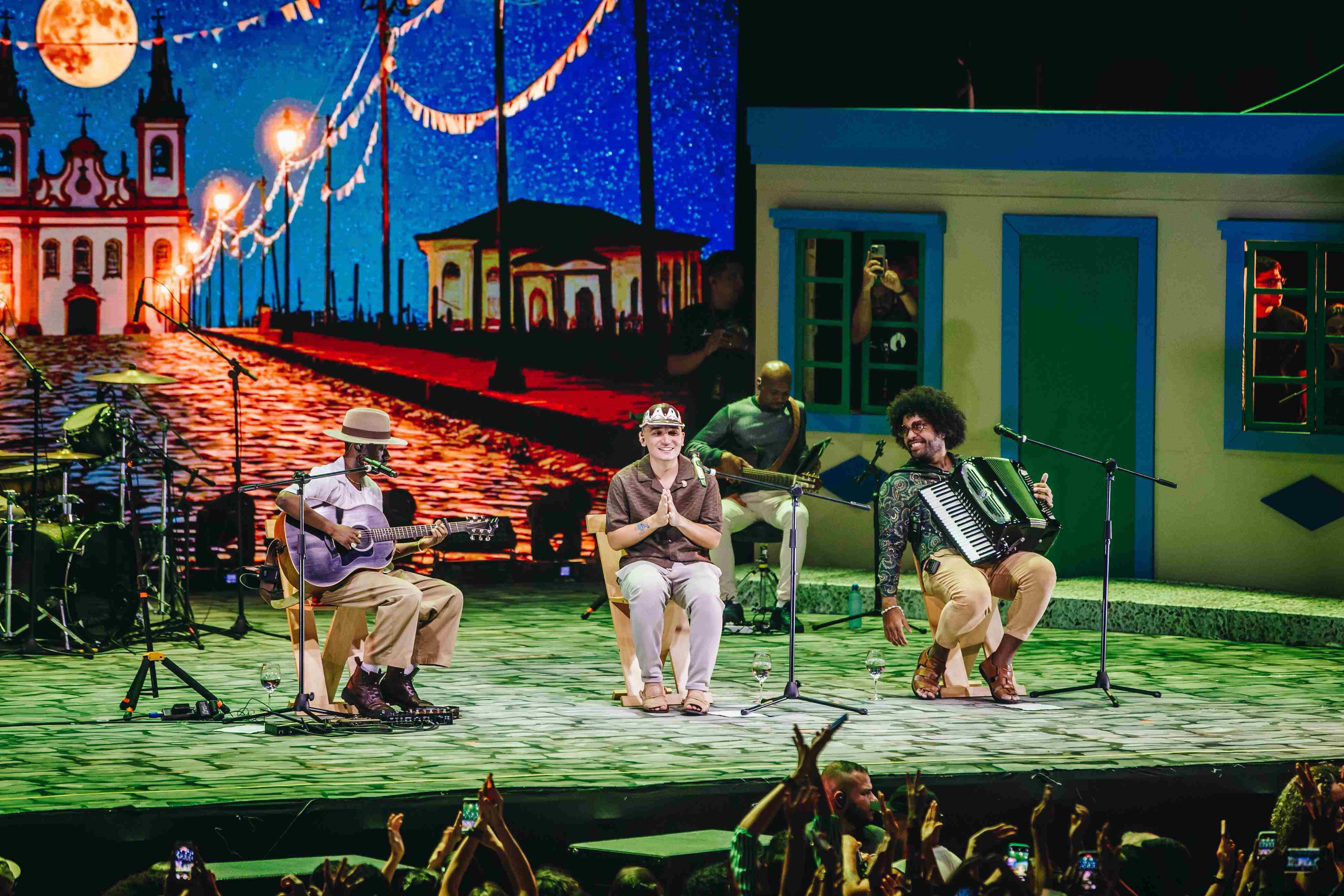 Show musical em um palco decorado com cenário que remete ao interior nordestino, com uma igreja iluminada sob a lua cheia ao fundo e uma casa colorida à direita. Três músicos estão sentados na frente: Jota.pê toca violão, João Gomes sorri com as mãos juntas, e Mestrinho toca sanfona. Ao fundo, outro músico toca baixo. O público aparece em frente ao palco, aplaudindo e registrando o momento com celulares.