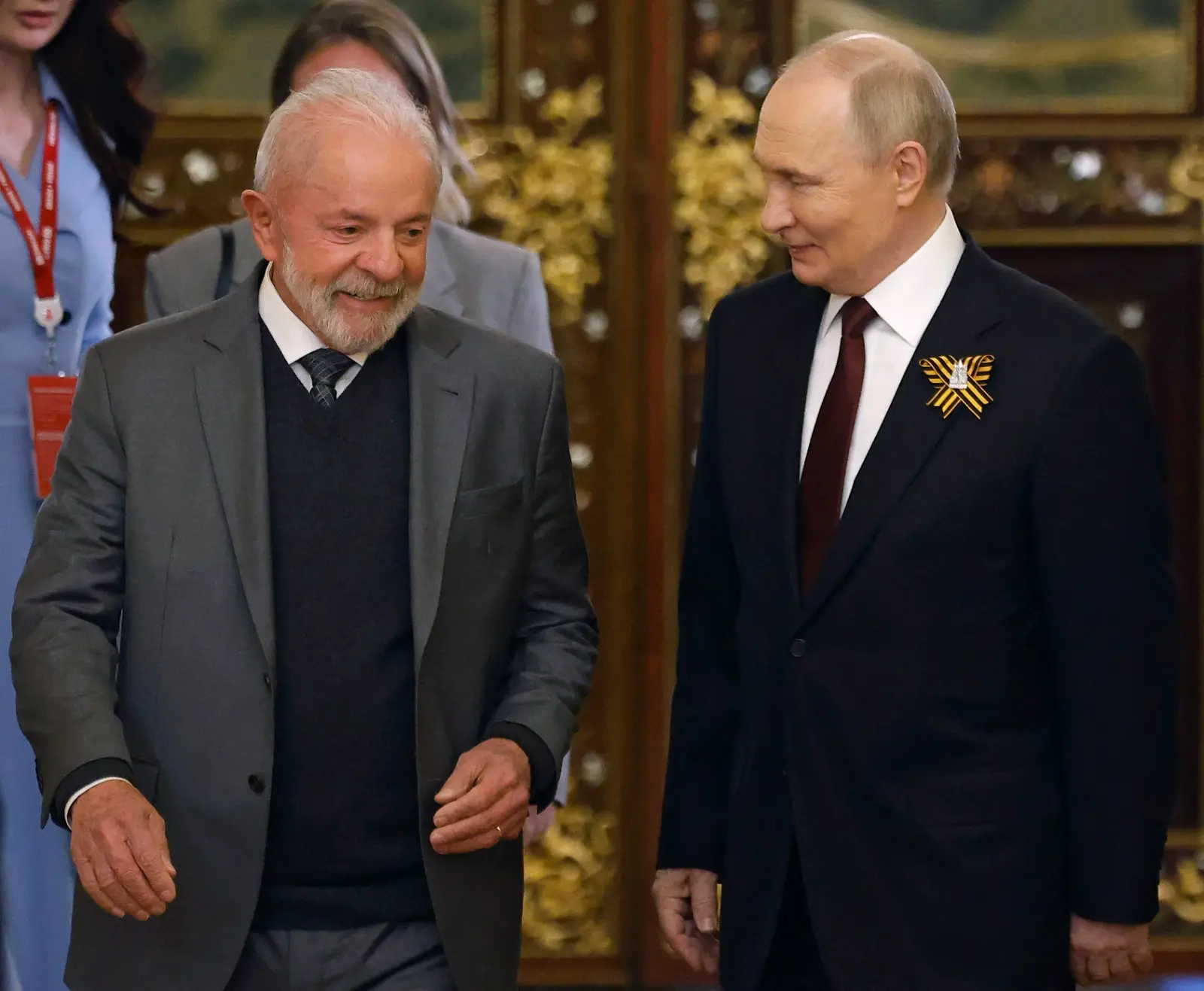 O presidente do Brasil, Luiz Inácio Lula da Silva, e o presidente da Rússia, Vladimir Putin, caminham lado a lado enquanto conversam sorrindo. Lula veste um terno cinza com suéter escuro por baixo, enquanto Putin usa um terno escuro com gravata bordô e carrega no paletó a fita de São Jorge, um símbolo militar russo. Ao fundo, há portas douradas ornamentadas e outras pessoas parcialmente visíveis.