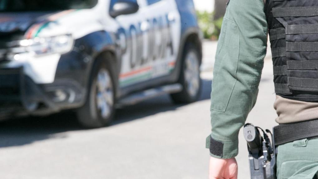 Ao fundo, uma viatura da polícia. Na parte direita da imagem, aparece o braço de um PM fardado