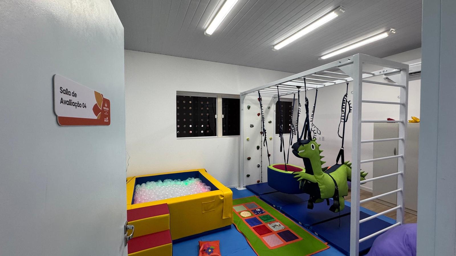 Imagem mostra sala de avaliação de crianças no Centro de Diagnóstico Espaço Girassol. O espaço tem uma piscina de bolinhas amarela, um dinossauro de pelúcia verde e tapetes coloridos no chão.