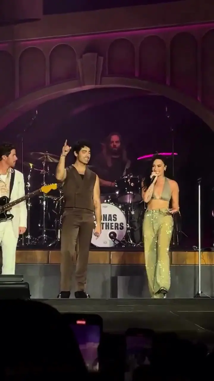 Apresentação de uma banda ao vivo em um palco, com dois vocalistas e músicos tocando instrumentos, cenário com iluminação rosa e fundo de palco.