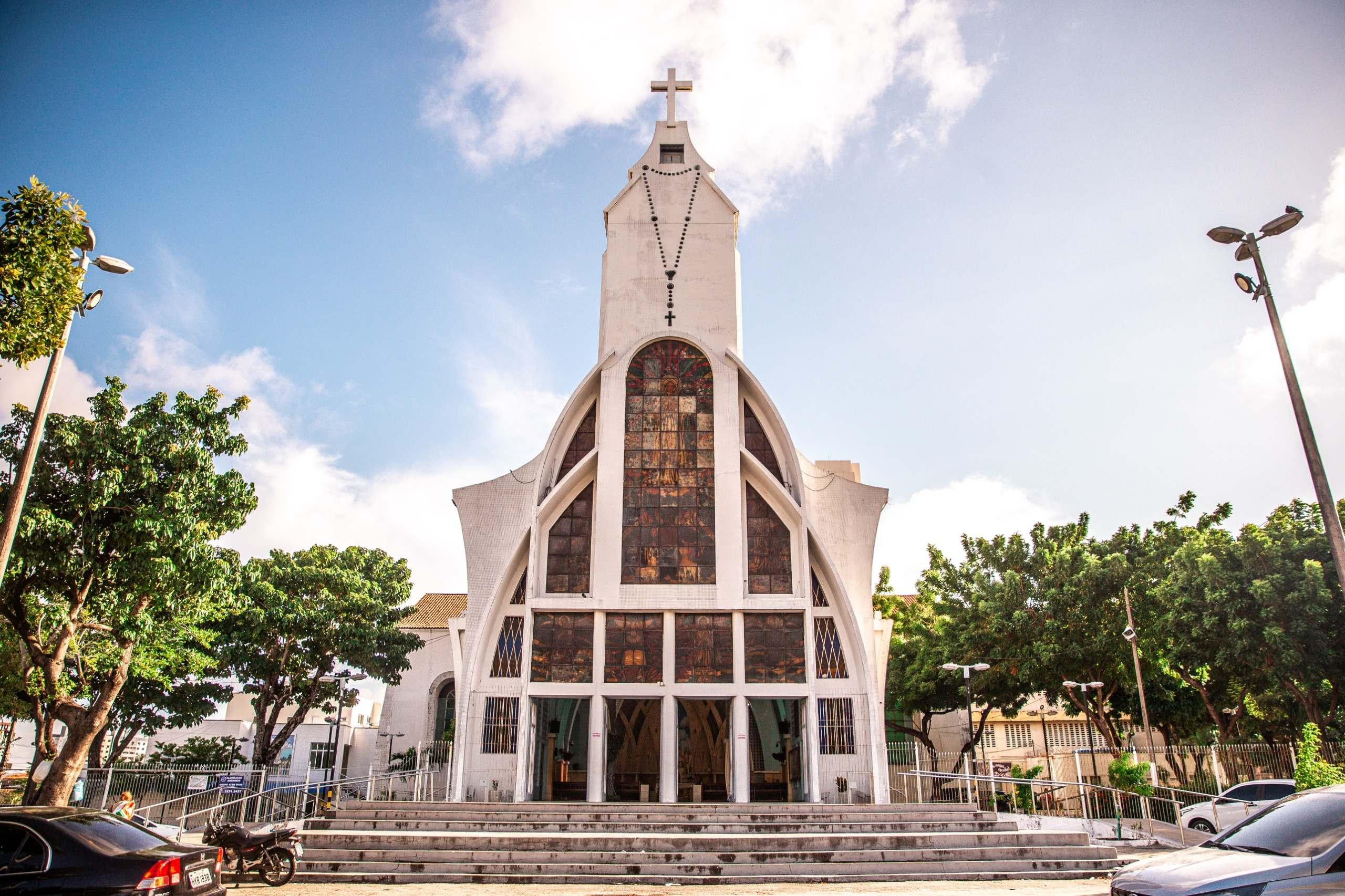Uma fotografia da fachada da Igreja de Fátima em Fortaleza, Ceará, vista de frente. O edifício é branco, com uma arquitetura moderna e curvilínea, e uma grande cruz no topo da torre sineira. Um rosário grande pendura da torre. A fachada é dominada por grandes vitrais em tons quentes. Uma escadaria leva à entrada principal da igreja. A imagem é enquadrada por árvores em ambos os lados e um céu azul com nuvens brancas.