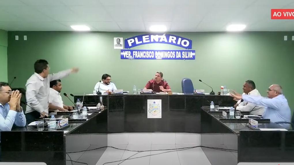 Imagem mostra trecho de sessão na Câmara Municipal de Palhano, com vereadores sentados em mesas dispostas em “U” e placa ao fundo com os dizeres “Plenário Ver. Francisco Domingos da Silva”. Alguns participantes gesticulam e discutem, indicando possível momento de debate acalorado. No canto superior direito, há um selo vermelho com o texto “AO VIVO”.