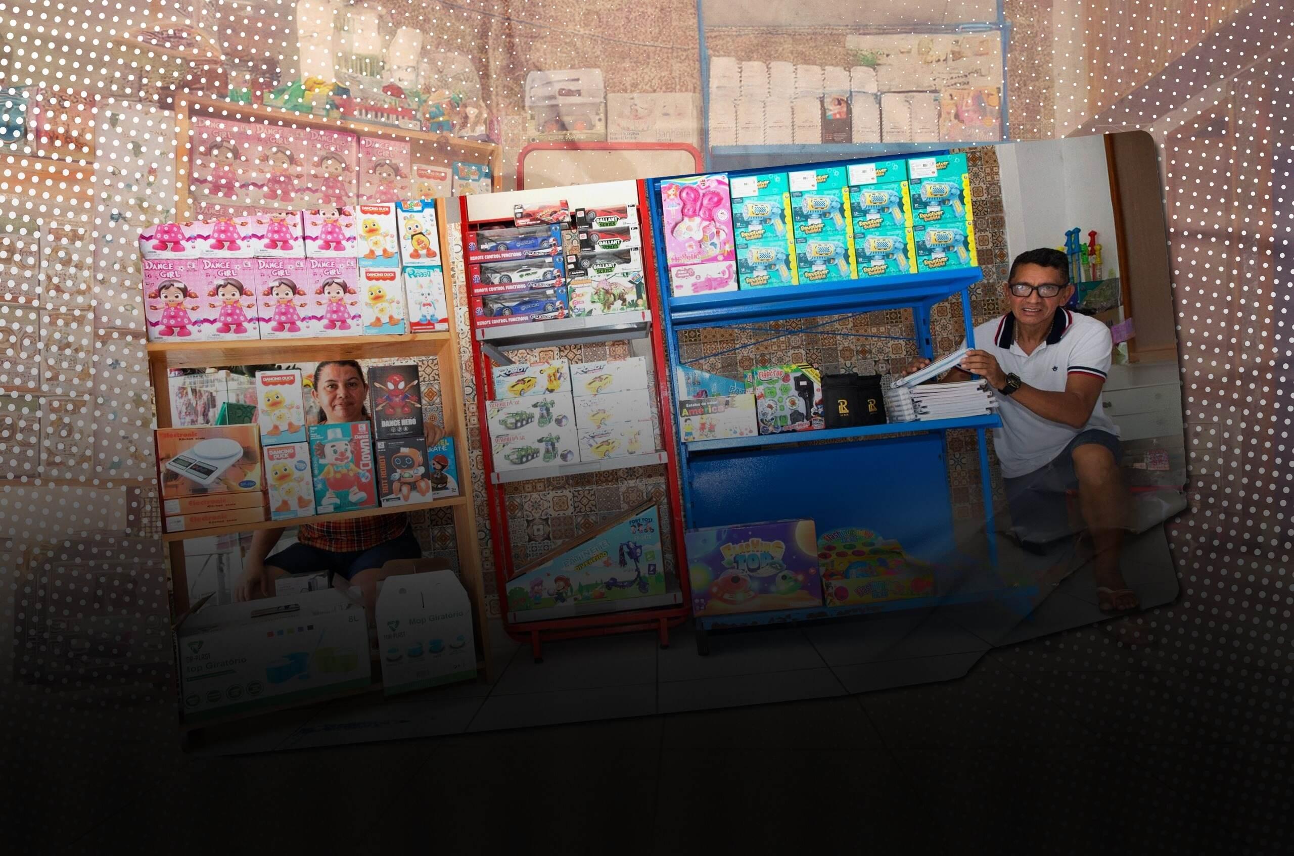 Dois comerciantes, um homem e uma mulher, posam sorrindo dentro de uma loja, ajoelhados ao lado de prateleiras repletas de brinquedos e utilidades domésticas. À esquerda, há caixas e bonecas, e à direita, jogos, pistolas de brinquedo e cadernos. O ambiente tem piso branco e parede com azulejos estampados ao fundo.