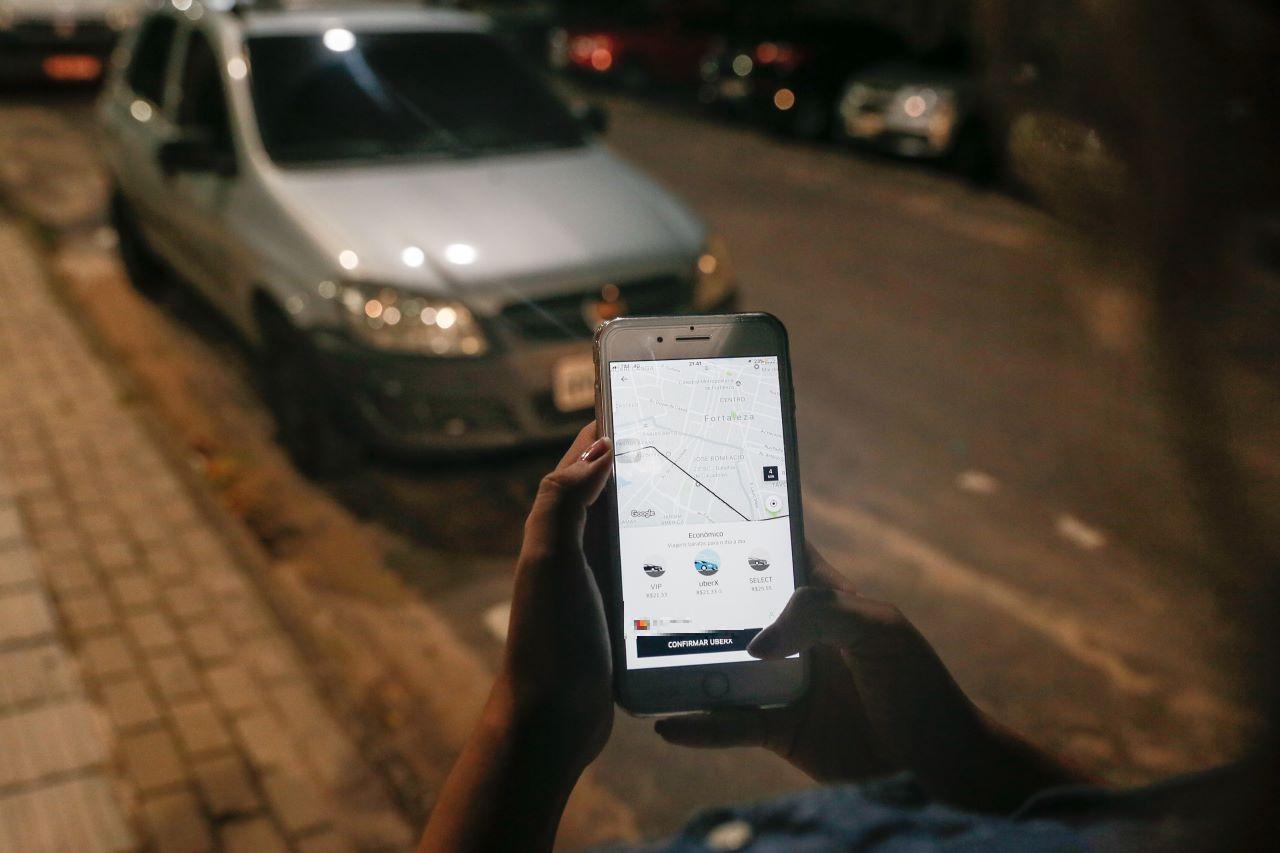 Pessoa usando smartphone para solicitar corrida na Uber em rua à noite, ilustrando operação da Polícia Civil do RJ que investiga quadrilha suspeita de usar inteligência artificial para fraudar Uber e gerar prejuízo de R$ 114 mil