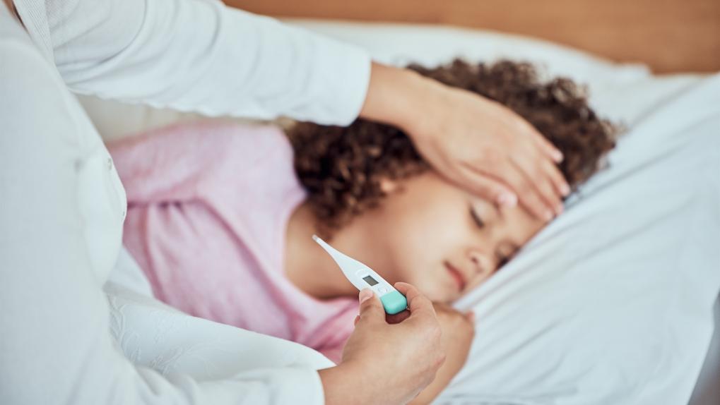 A foto mostra uma criança dormindo em uma cama enquanto um adulto toca sua testa e segura um termômetro digital na outra mão.