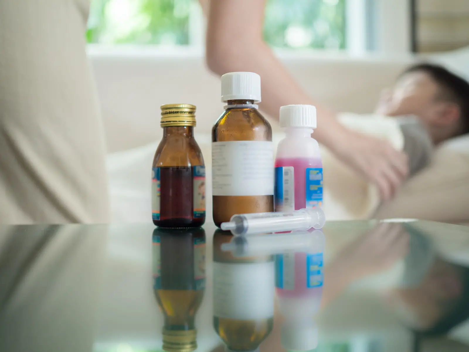 Imagem mostra três frascos de remédio e uma seringa para dosagem sobre uma mesa de vidro. Ao fundo, uma criança deitada é visível de forma desfocada.