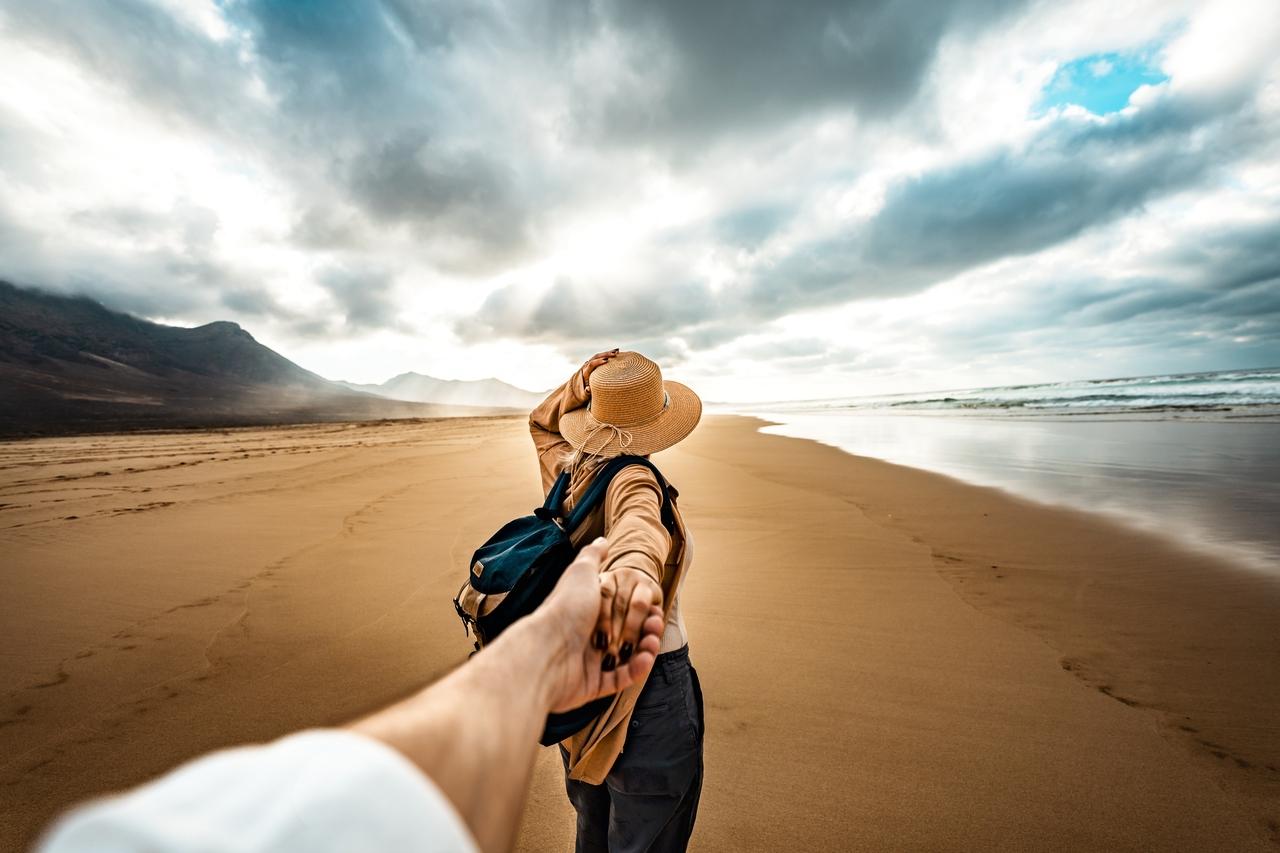 Cena de uma mulher de chapéu de palha segurando a mão de quem a fotografa enquanto caminha na praia sob céu nublado, clima de aventura e conexão com a natureza, ilustrando semana mais barata para viajar que acontece em agosto