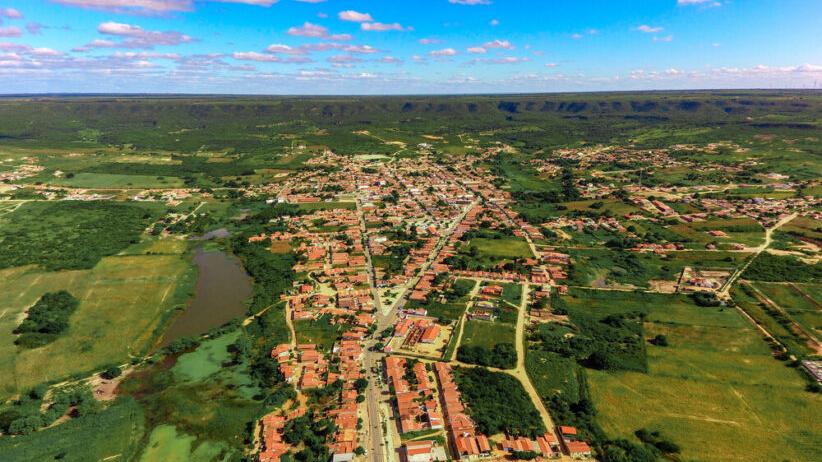 Imagem aérea de uma cidade pequena rodeada por áreas verdes e campos, com ruas principais alinhadas e casas de telhados avermelhados. À esquerda, há um lago ou açude cercado por vegetação. Ao fundo, observa-se uma serra extensa no horizonte, sob um céu azul com algumas nuvens espalhadas.