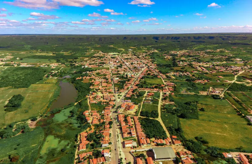 Imagem aérea de uma cidade pequena rodeada por áreas verdes e campos, com ruas principais alinhadas e casas de telhados avermelhados. À esquerda, há um lago ou açude cercado por vegetação. Ao fundo, observa-se uma serra extensa no horizonte, sob um céu azul com algumas nuvens espalhadas.