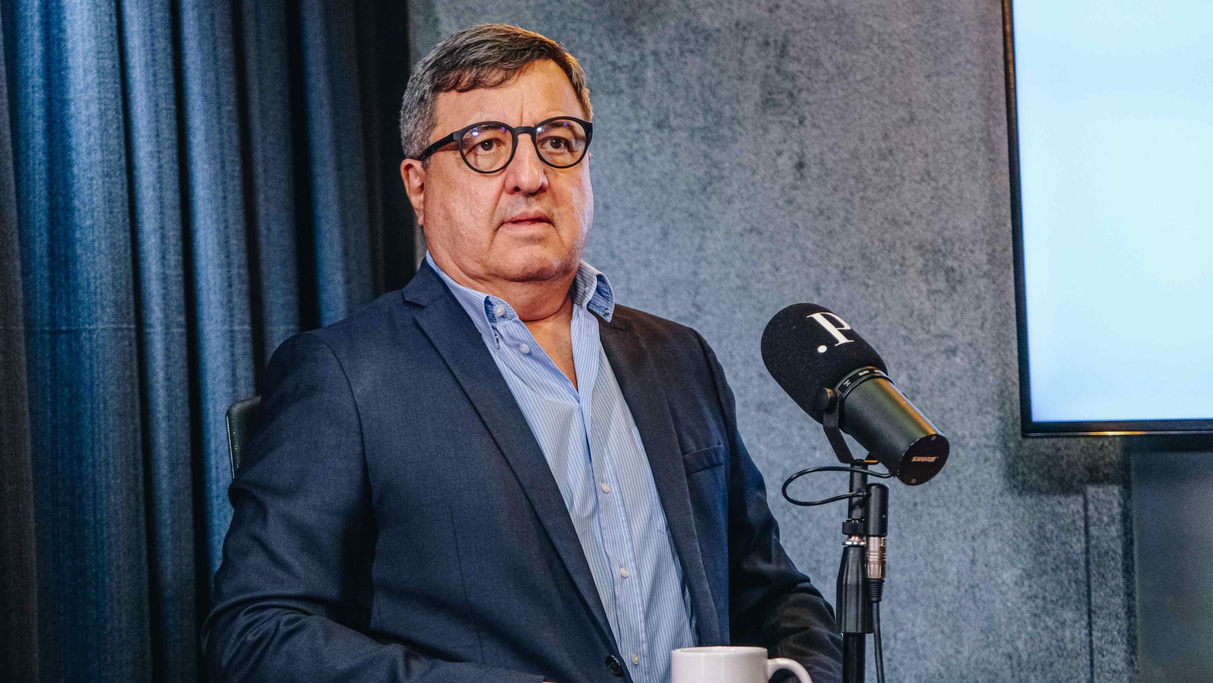 Na imagem, o deputado federal Danilo Forte (União) aparece sentado em um estúdio de gravação, vestindo paletó azul-escuro e camisa social azul-clara, sem gravata. Ele usa óculos de armação escura e mantém uma expressão séria enquanto fala ao microfone, que exibe a letra “P” — símbolo do programa PontoPoder. À frente dele, há uma caneca branca com o mesmo logotipo. Ao fundo, cortinas escuras e uma tela iluminada com a palavra “PONTO” compõem o cenário.