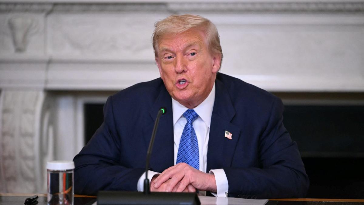 Donaldo Trump de terno azul-marinho e gravata azul falando em um microfone em uma sala com decoração clássica branca. Ele está sentado à mesa com as mãos cruzadas e um copo de água à sua esquerda. Uma bandeira dos EUA está presa na lapela do paletó.