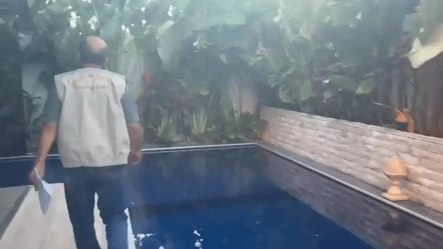 Homem de costas caminhando ao lado de uma piscina em área externa cercada por plantas tropicais. Ele veste colete bege com inscrições nas costas e segura papéis na mão direita. À direita da piscina, há um muro de pedras com luminárias decorativas. A cena parece estar relacionada a uma inspeção ou operação oficial.
