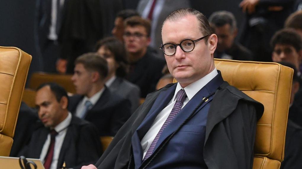 Zanin é um homem de terno azul, gravata roxa e toga preta, sentado em uma cadeira de couro marrom, com expressão séria. Ele usa óculos redondos e está em um ambiente formal, no próprio STF, com outras pessoas ao fundo, também vestidas formalmente.