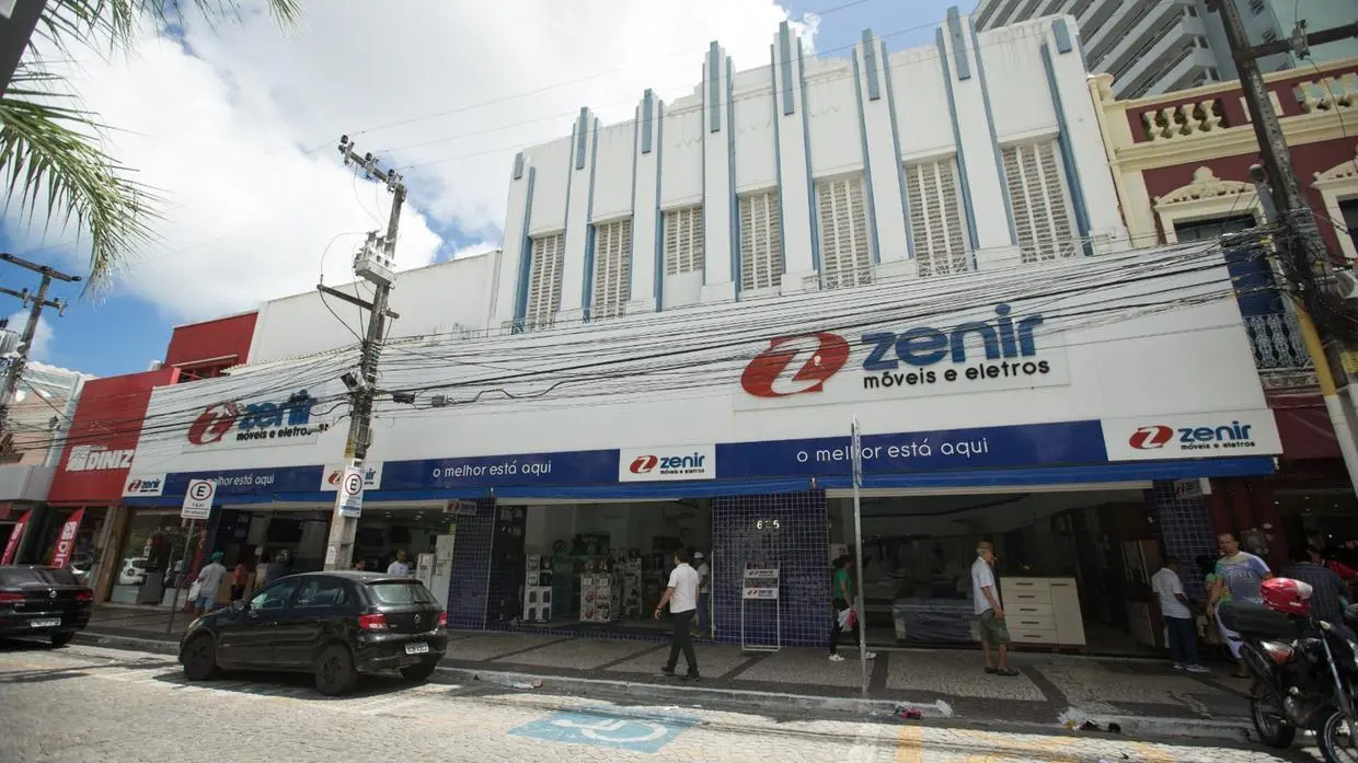A imagem mostra a loja da Zenir Móveis e Eletros em um prédio de dois andares com fachada branca e detalhes em azul-claro, localizada em uma rua movimentada de Fortaleza