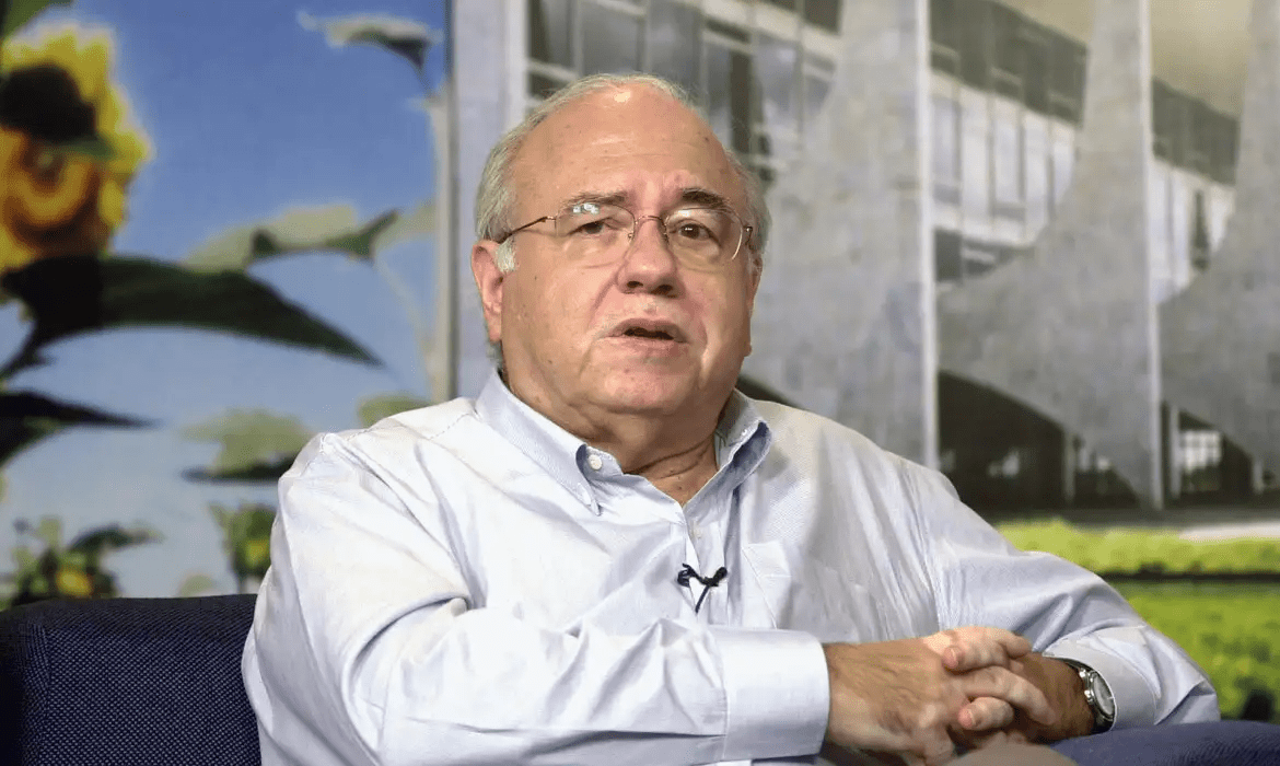 Luis Fernando Veríssimo é um homem idoso de cabelos grisalhos e óculos, vestindo camisa social clara, sentado com as mãos cruzadas. Ao fundo, há uma imagem com flores e uma estrutura arquitetônica moderna, provavelmente em um estúdio ou ambiente de entrevista.