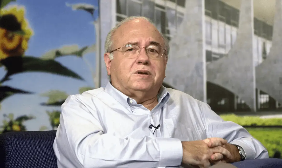 Luis Fernando Veríssimo é um homem idoso de cabelos grisalhos e óculos, vestindo camisa social clara, sentado com as mãos cruzadas. Ao fundo, há uma imagem com flores e uma estrutura arquitetônica moderna, provavelmente em um estúdio ou ambiente de entrevista.