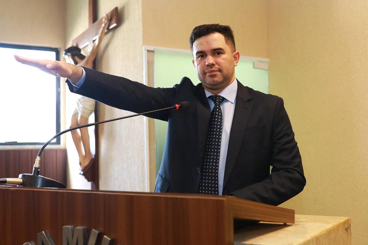 Homem de terno escuro e gravata azul com bolinhas fala ao microfone em um púlpito. Ao fundo, há bandeiras do Brasil, do estado do Ceará e do município de Fortaleza, além de parte de um letreiro na parede, indicando que se trata do auditório da Câmara de Fortaleza. A cena ocorre em um ambiente formal.