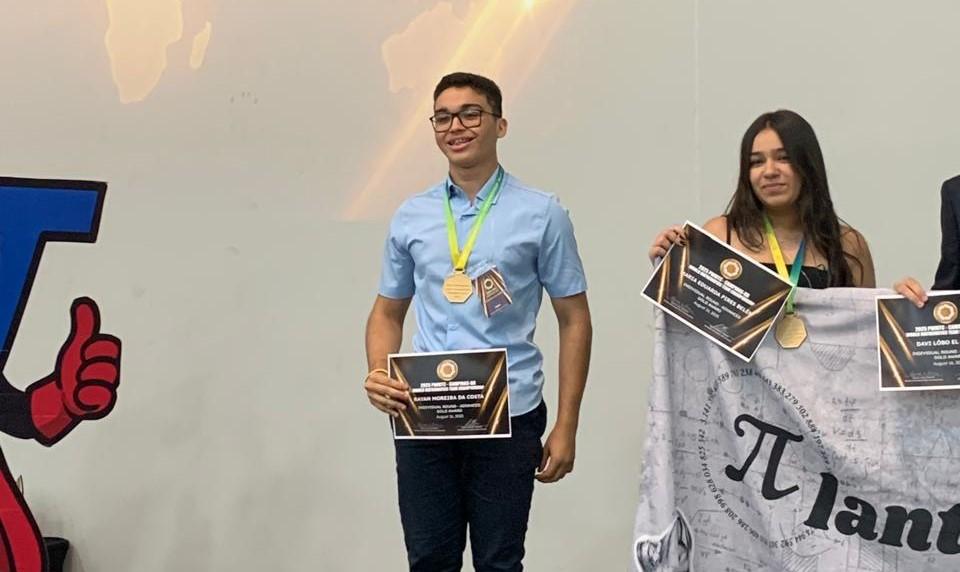 Um jovem de óculos e camisa azul, no centro, segura um certificado de premiação dourado e usa duas medalhas douradas no pescoço. Ele sorri enquanto está no palco. À sua direita, uma jovem de cabelos escuros e blusa preta também segura um certificado e uma medalha dourada. Ao fundo, um telão mostra um mapa-múndi, e uma faixa com o símbolo do Pi e a palavra 