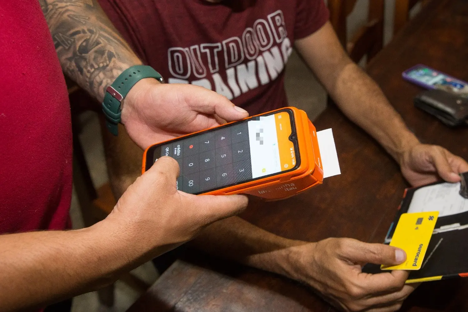Foto que contém homem segurando maquineta para pagamento de conta, enquanto homem se prepara com o cartão em mãos para efetuar o pagamento