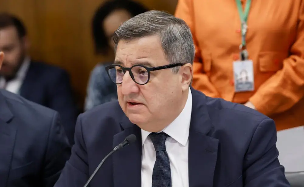 Um homem de meia-idade, de terno azul-escuro, camisa branca e gravata preta, fala ao microfone em um ambiente formal, como uma audiência ou reunião oficial. Ele usa óculos de aro preto e tem cabelos curtos grisalhos. Ao fundo, aparecem pessoas desfocadas, incluindo uma mulher em pé com roupa laranja e crachá no pescoço.