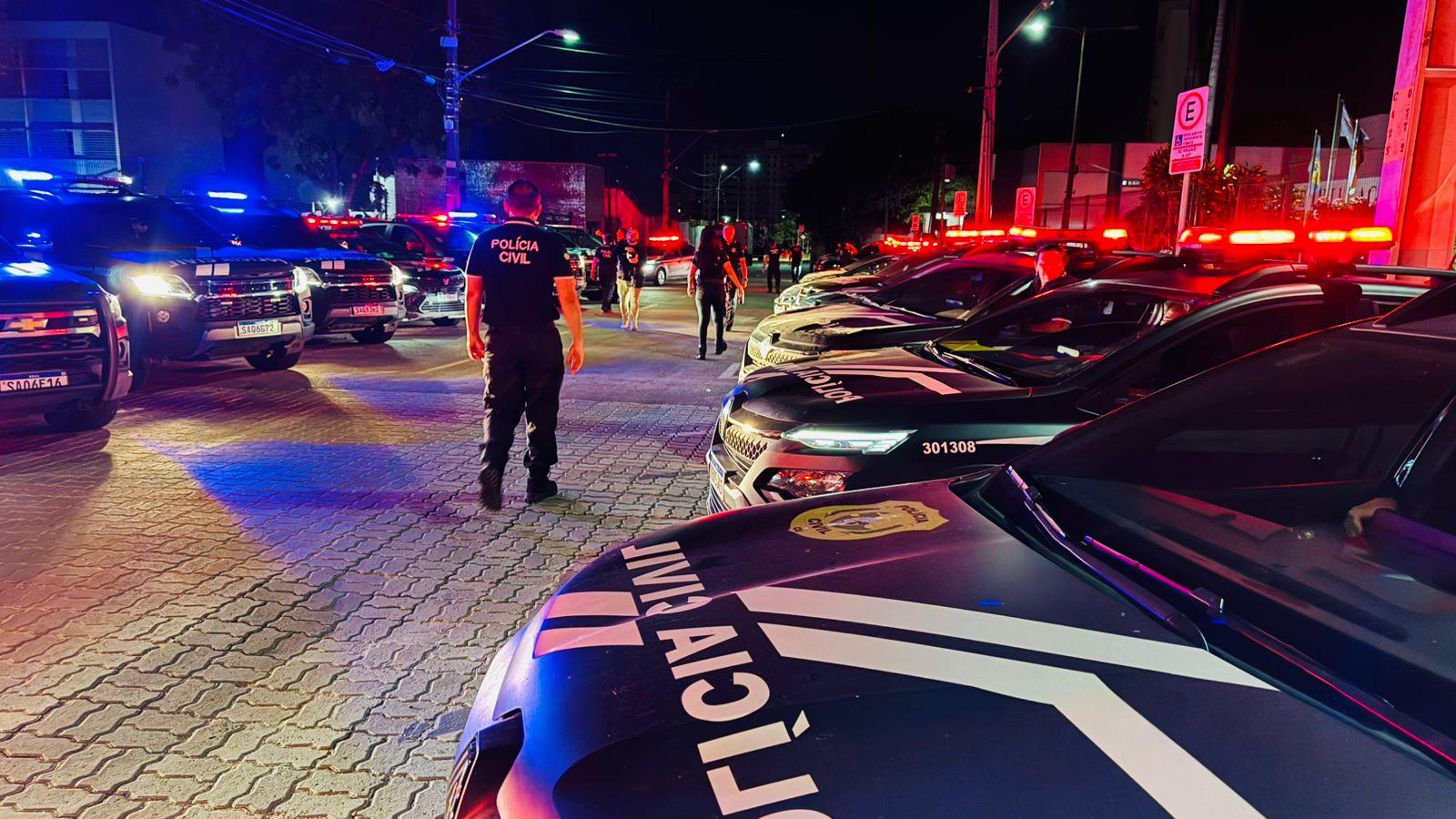 Câmeras de segurança mostram uma cena de emergência com várias viaturas policiais e policiais civis em uma operação noturna na rua, com iluminação de sinais de emergência.