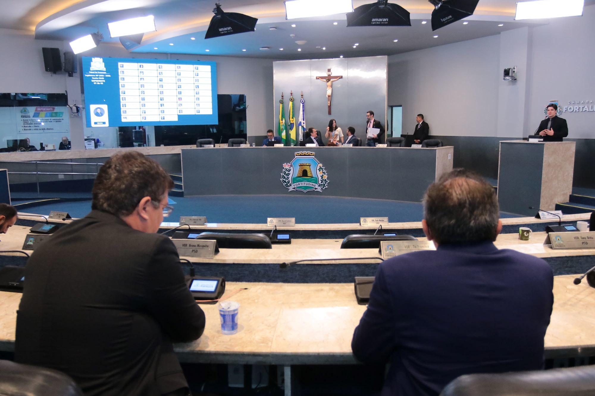 Sessão na Câmara Municipal de Fortaleza. Vereadores estão sentados no plenário, olhando para o painel eletrônico que exibe a lista de presença e votação. Ao fundo, na mesa diretora, estão autoridades e servidores. Na parede central, o brasão de Fortaleza, bandeiras do Brasil, do Ceará e de Fortaleza, e um crucifixo