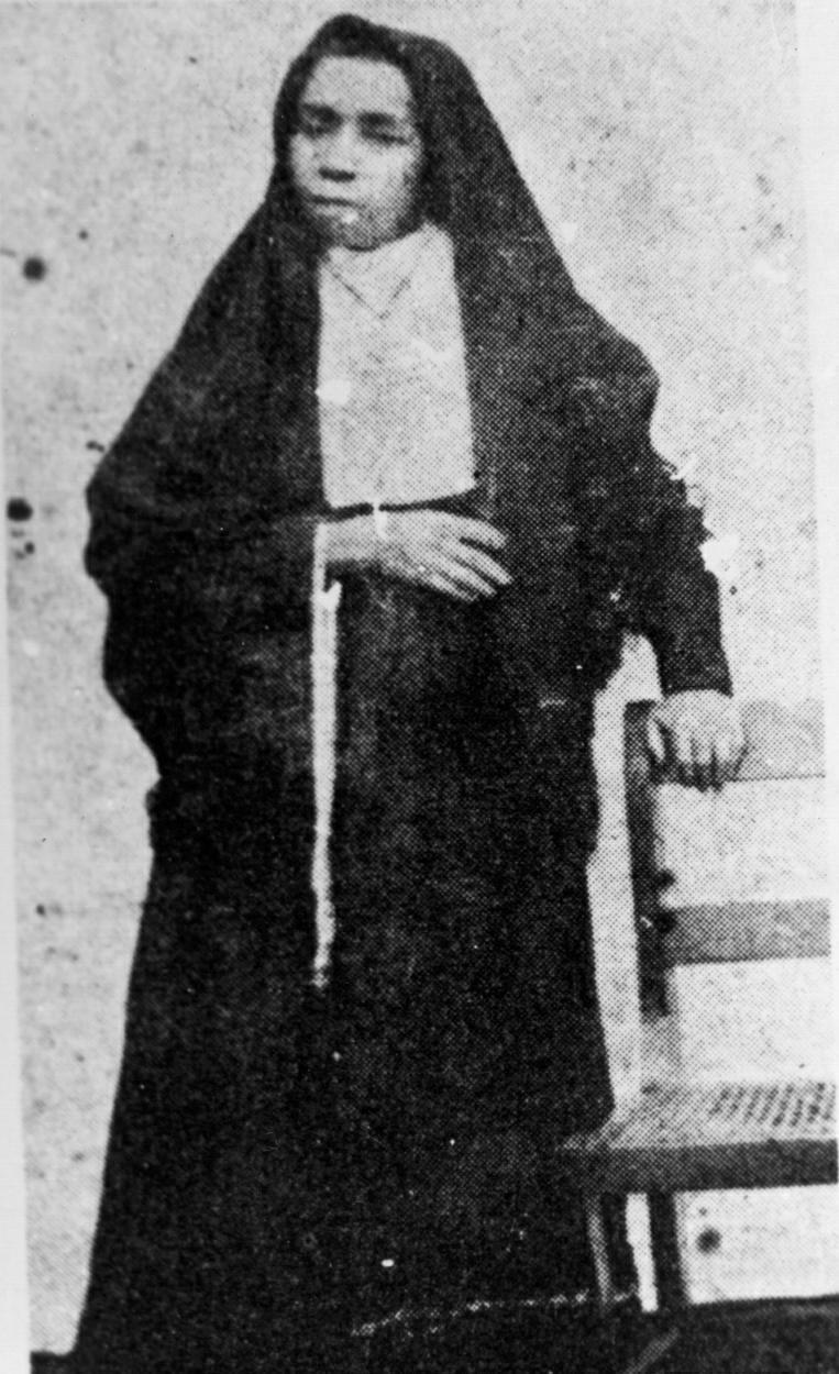 A foto em preto e branco antiga mostra uma mulher com veste de beata, um véu preto na cabeça e apoiada com o braço esquerdo em uma cadeira. Seu semblante é sério.