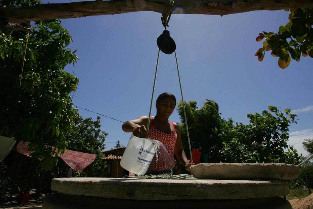 Foto de mulher puxando água de poço com corda. Ao fundo, céu claro, árvores e roupas no varal