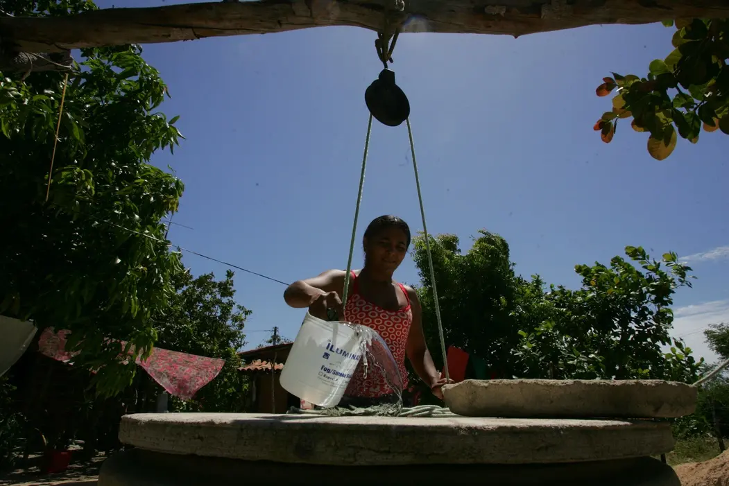 Foto de mulher puxando água de poço com corda. Ao fundo, céu claro, árvores e roupas no varal