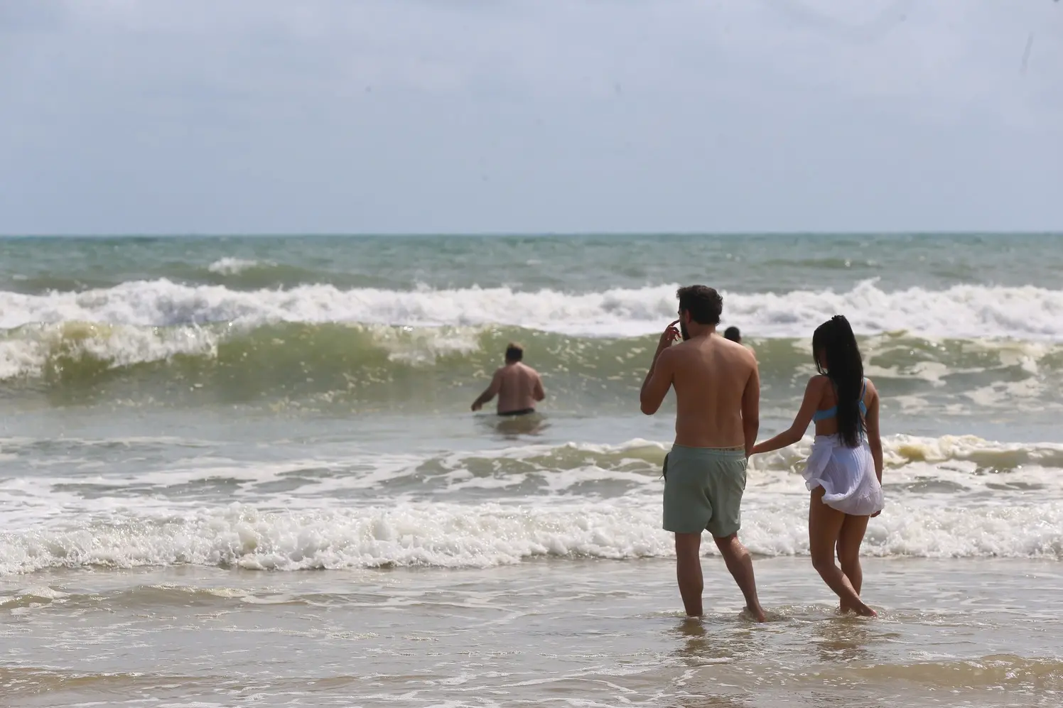 Foto mostra banhistas aproveitando o mar na Praia do Futuro, com ondas fortes e céu parcialmente nublado. Em primeiro plano, um casal heterossexual caminha pela água rasa de mãos dadas.