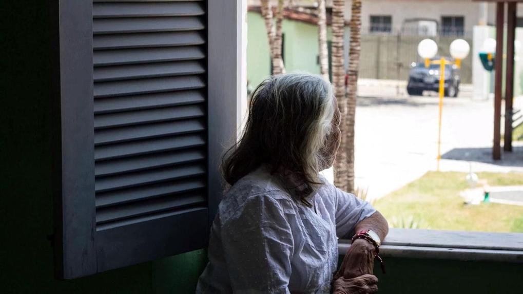 Mulher idosa de cabelos grisalhos, vestindo blusa branca, está encostada na janela e olha para fora, em direção à rua iluminada pelo sol.