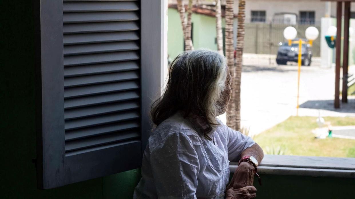 Mulher idosa de cabelos grisalhos, vestindo blusa branca, está encostada na janela e olha para fora, em direção à rua iluminada pelo sol.
