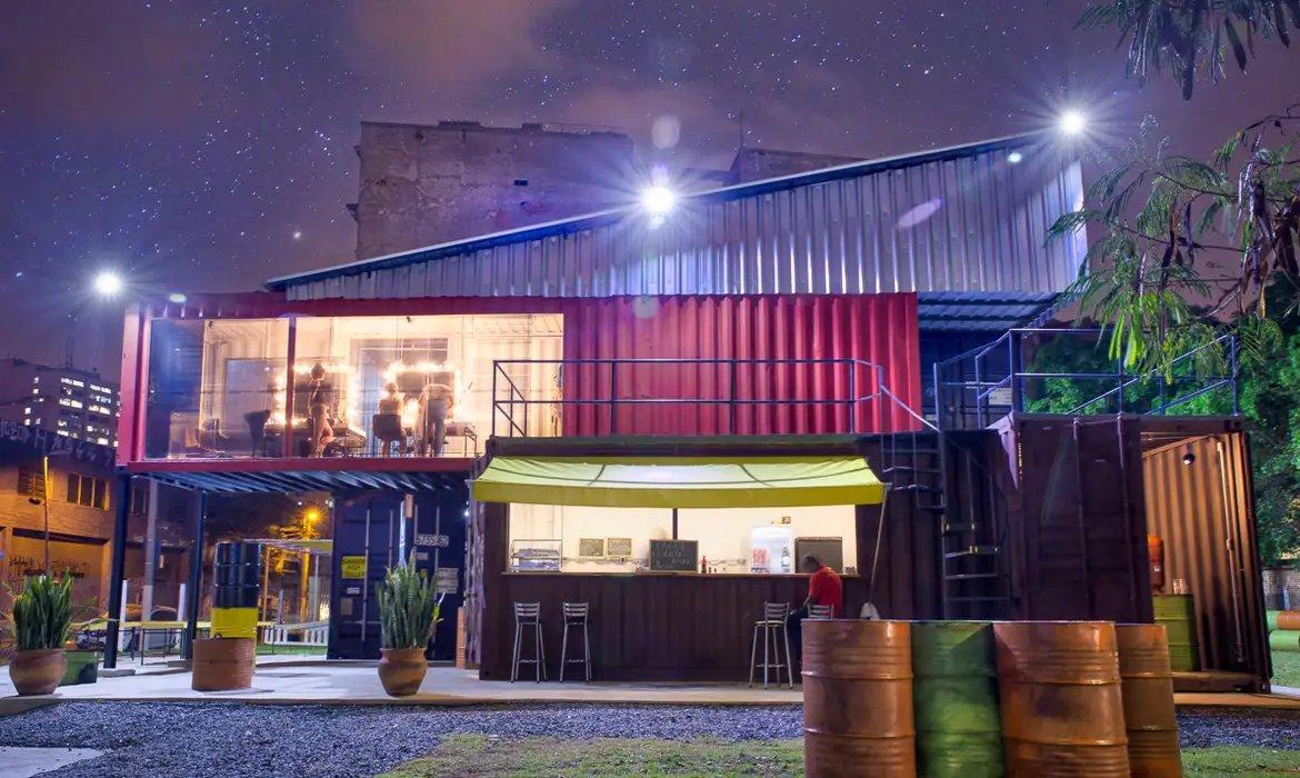 Imagem de um restaurante em container com ambiente ao ar livre iluminado à noite, com céu estrelado ao fundo, integrando design moderno e sustentável.