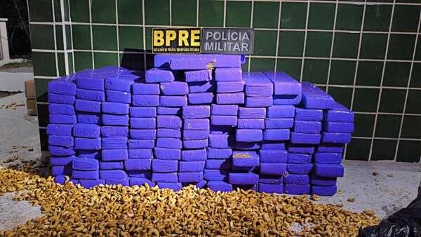 Pacotes roxos de maconha empilhados com castanhas espalhadas no chão e placas da Polícia Militar e BPRE acima.