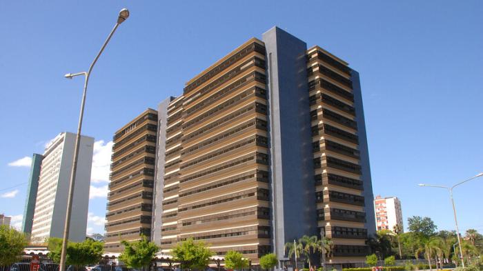 Imagem de um edifício alto e moderno em uma cidade, com céu claro ao fundo, árvores ao redor e uma rua na frente, destacando-se na paisagem urbana.