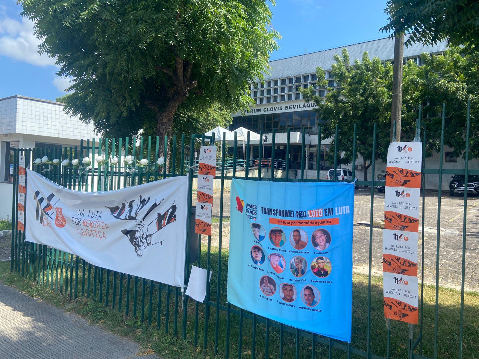 Banners e faixas de protesto contra a violência e a injustiça, exibidas em uma grade de ferro na rua, com árvores ao fundo e um prédio ao fundo em uma manhã ensolarada