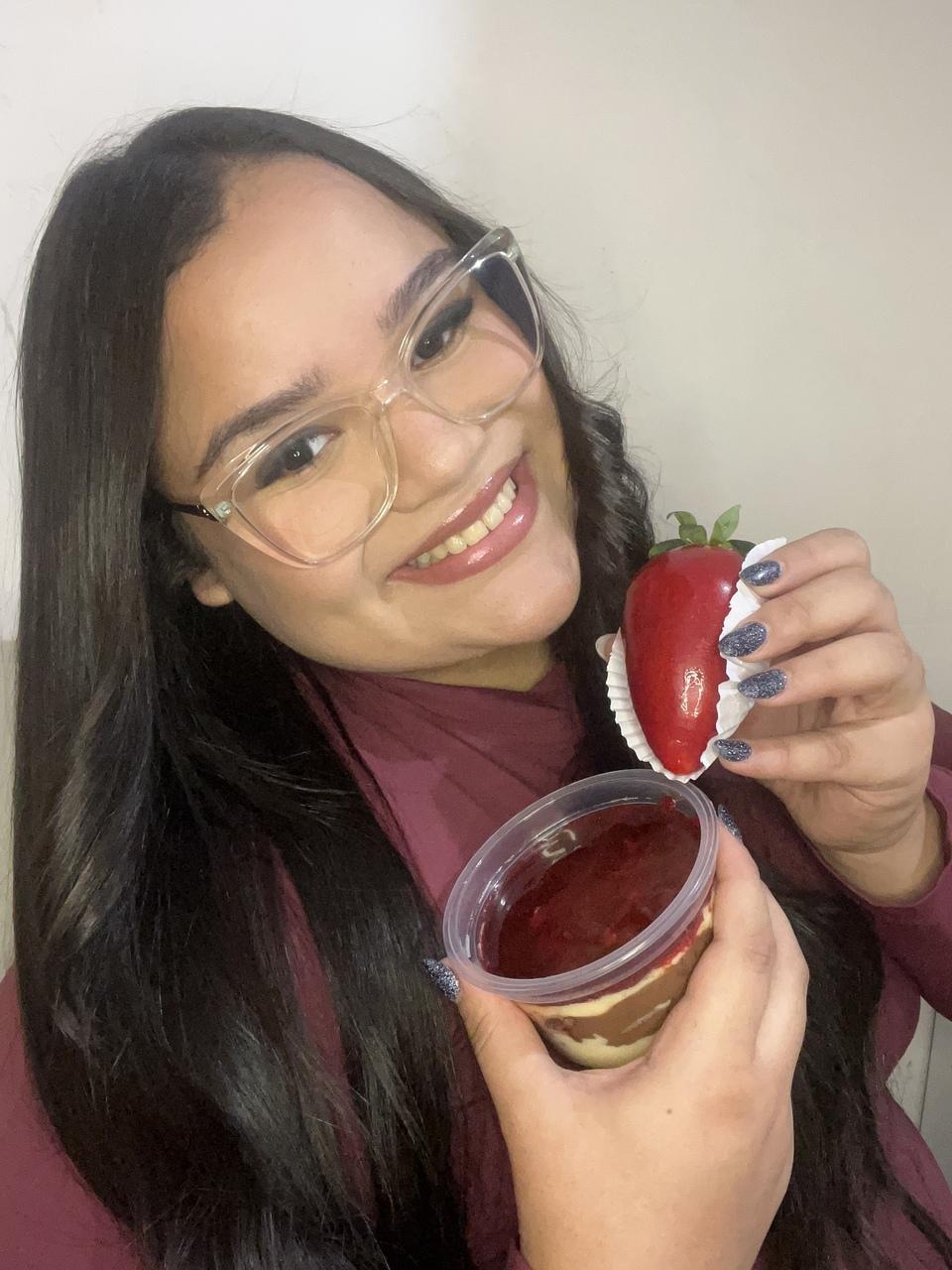 Imagem de Sabrina Leite segurando um doce morango do amor e um pote com doce de morangos