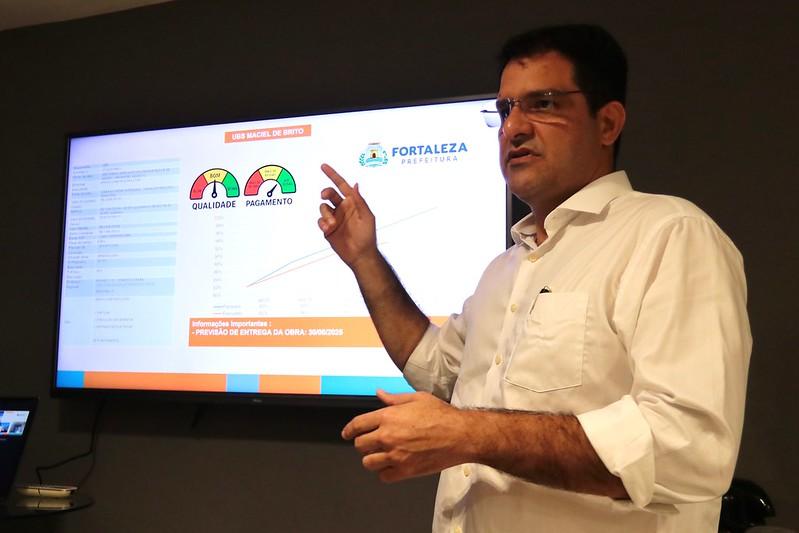 Homem de óculos e camisa branca apresenta dados em uma tela, apontando para gráficos com indicadores de qualidade e pagamento de obras públicas da Prefeitura de Fortaleza.