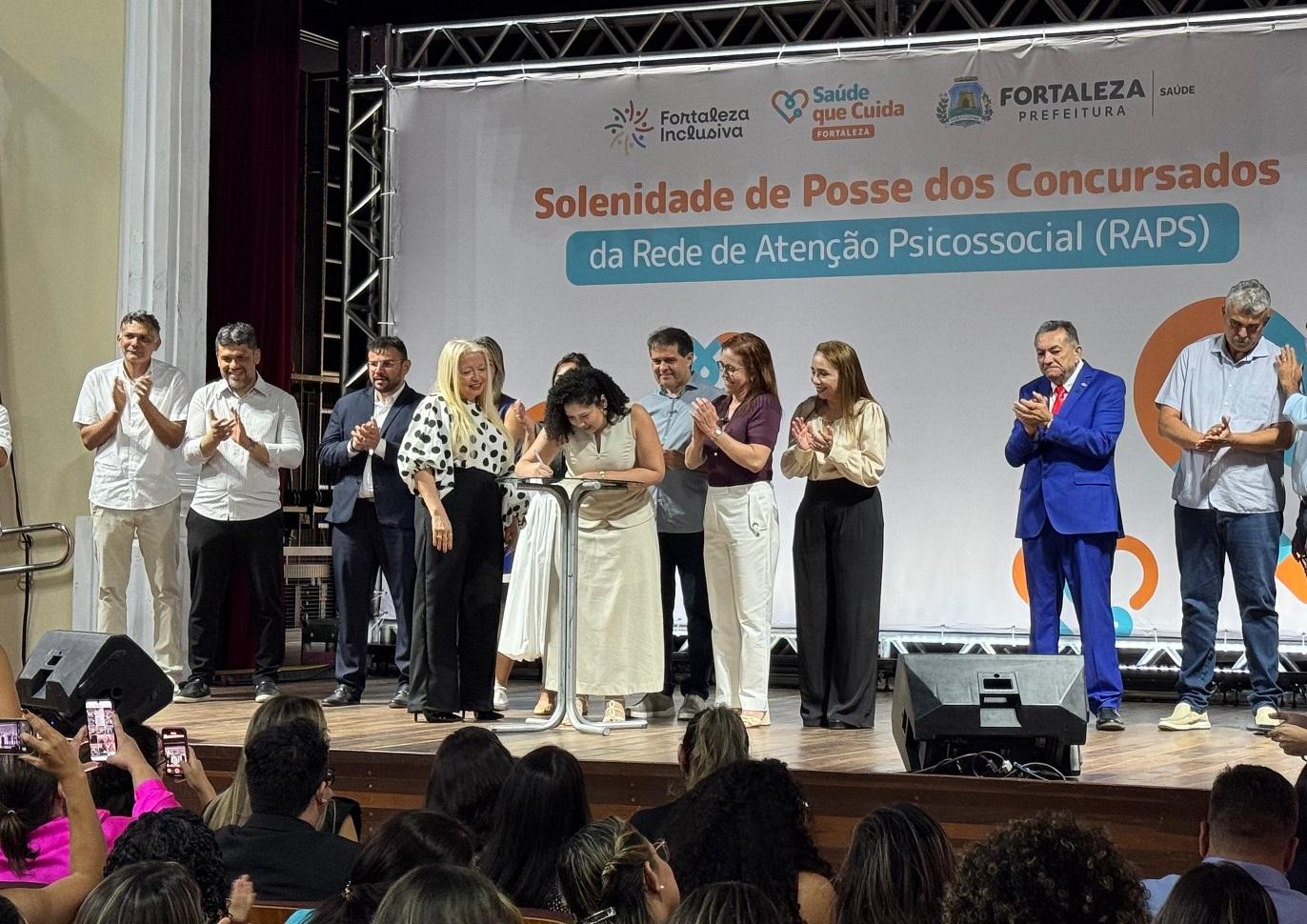Foto mostra a solenidade de posse dos concursados da Rede de Atenção Psicossocial (RAPS) em Fortaleza, com autoridades e novos servidores no palco. Uma mulher assina um documento no centro, enquanto os demais aplaudem e a plateia registra o momento.
