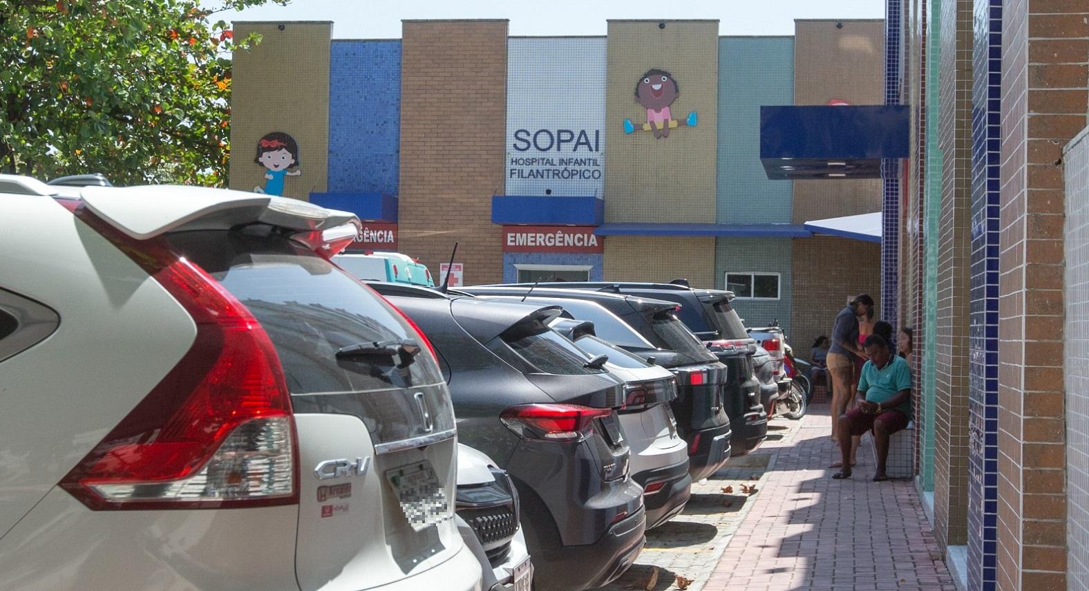 Foto mostra a entrada do Hospital Infantil Filantrópico Sopai, em Fortaleza, com a placa de emergência visível. Vários carros estão estacionados em frente e algumas pessoas aguardam do lado de fora do prédio.
