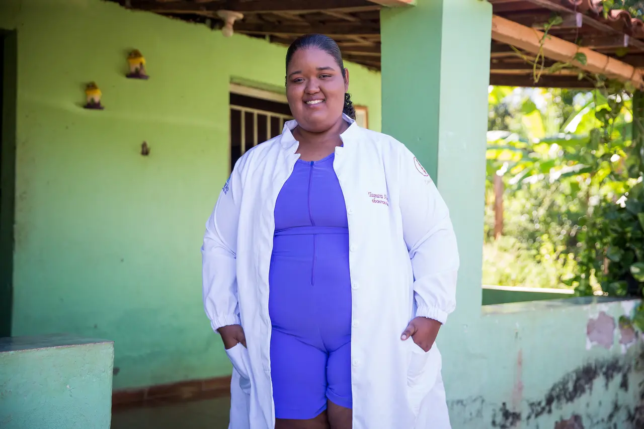 Taynara Alves de Almeida, mulher negra, sorrindo com roupa azul e um jaleco branco sobreposto