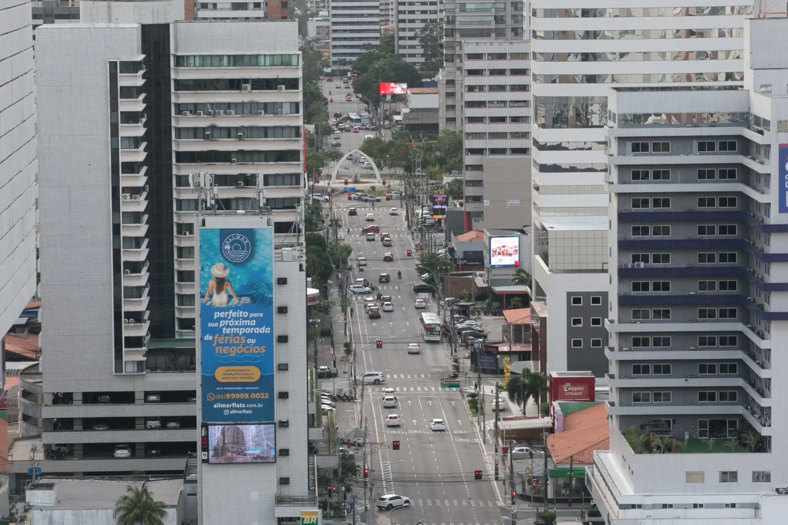Vista urbana mostrando uma rua movimentada ladeada por prédios altos em ambos os lados. A rua tem vários carros e múltiplas faixas. Há anúncios em alguns prédios, incluindo um grande em azul e amarelo à esquerda que diz 'perfeito para passar uma temporada de férias ou negócios'. Ao fundo, há mais edifícios altos e áreas verdes