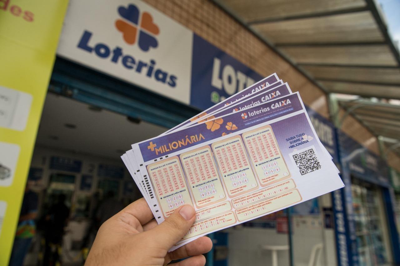 Imagem mostra mão masculina segurando bilhetes da loteria +milionária, com casa lotérica ao fundo, ilustrando loterias da caixa com sorteio neste dia.