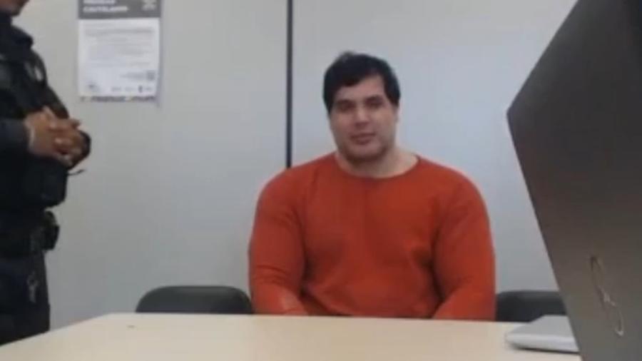 Homem sentado em uma mesa de escritório, com fundo de parede branca e uma pessoa de braços cruzados ao lado, em ambiente corporativo.