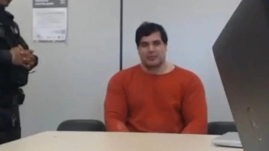 Homem sentado em uma mesa de escritório, com fundo de parede branca e uma pessoa de braços cruzados ao lado, em ambiente corporativo.