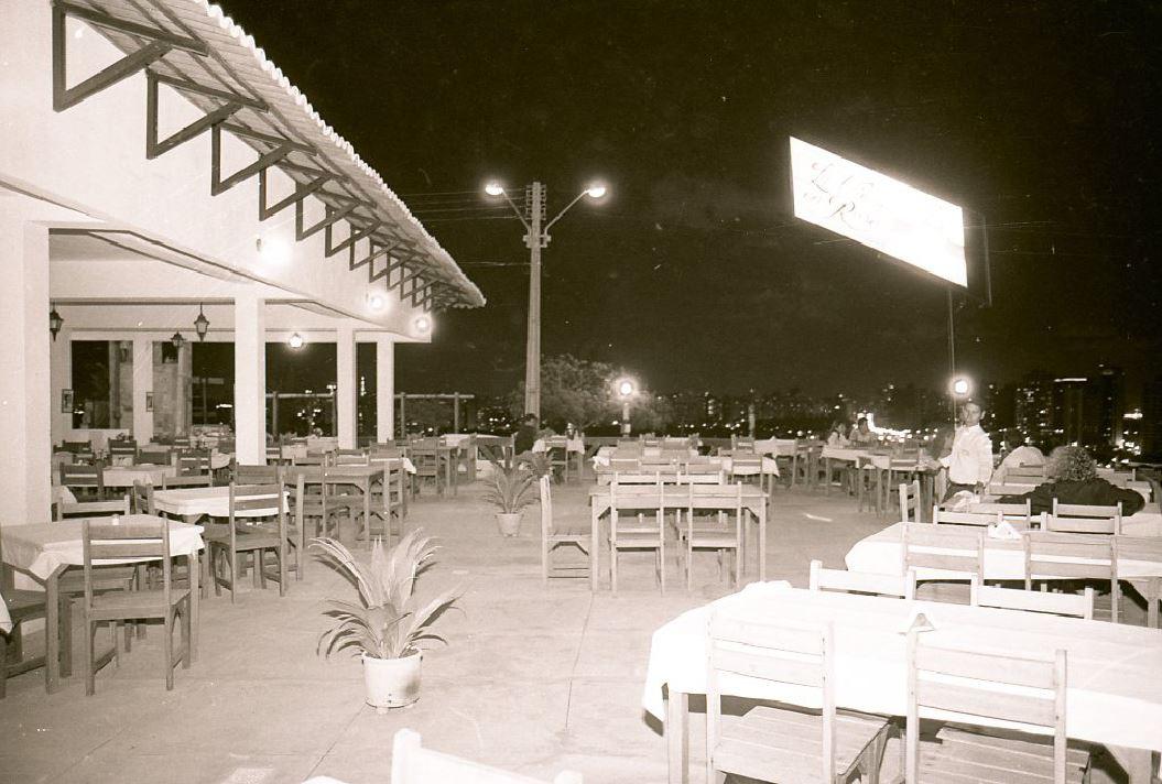 Foto antiga mostra um restaurante ao ar livre, com várias mesas cobertas por toalhas claras e poucas pessoas presentes. Ao fundo, vê-se a cidade iluminada à noite, criando um contraste com o espaço mais vazio em primeiro plano.