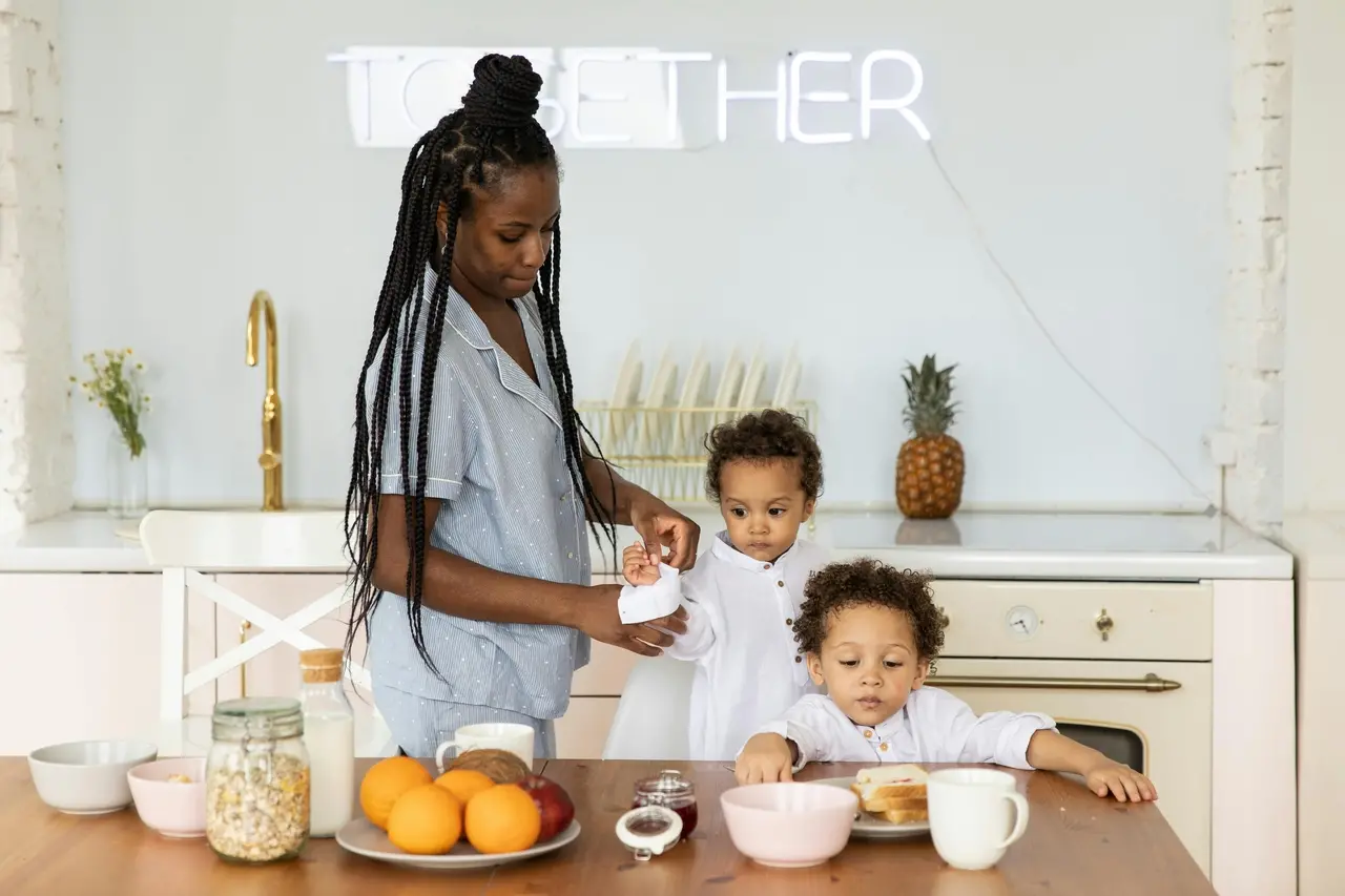 Mulher em uma cozinha ajudando duas crianças pequenas sentadas à mesa, com frutas, pão e leite sobre a mesa.