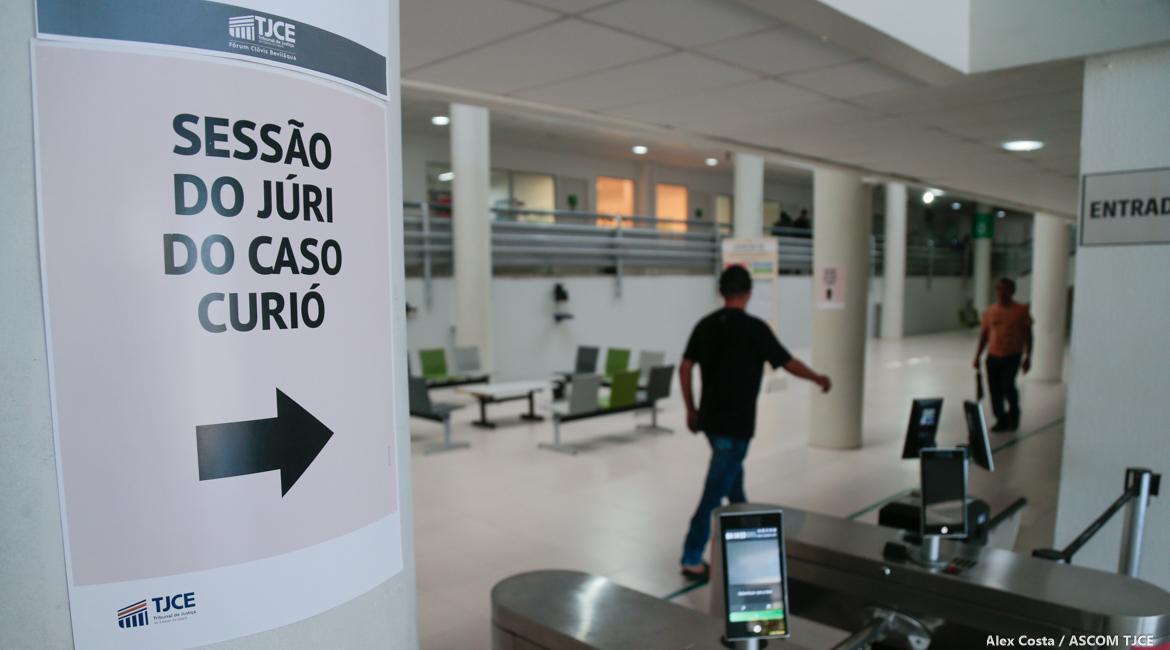Pilar com cartaz no TJ-CE e dois homens ao fundo, desfocados e andando. Imagem usada em matéria sobre o depoimento de PM que nega ao júri participação na Chacina do Curió